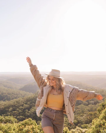 Woman in casual outfit stands with arms outstretched amidst a lush green forest backdrop, bathed in soft sunlight, creating a serene scene.
