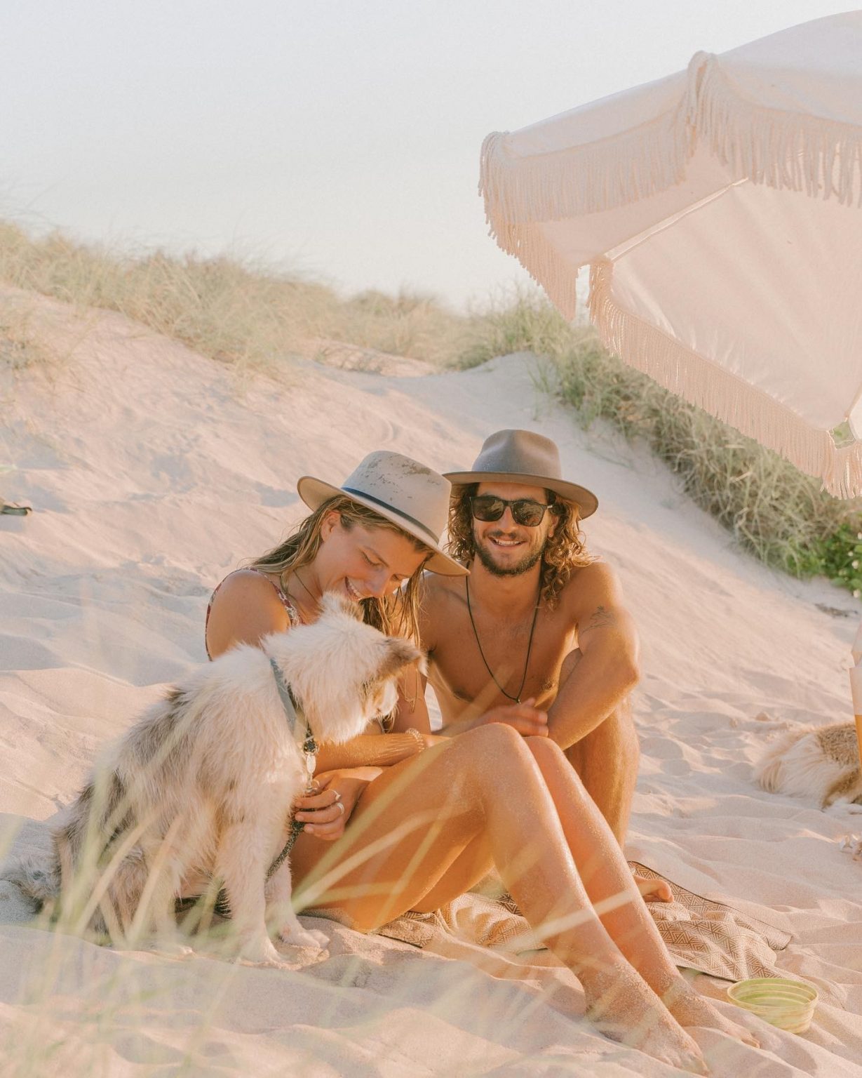 Couple sitting on a sand dune with a dog and parasol. The man and woman both wear hats and smile, illuminated by soft, natural light.