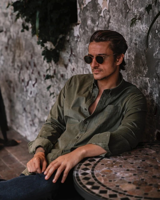 A man with brown hair and sunglasses is posing next to a stone table and plaster wall wearing an unbuttoned green collared shirt.