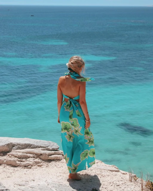 A woman in a tropical-print dress stands on a rocky cliff overlooking a turquoise ocean, enjoying the serene coastal scenery.
