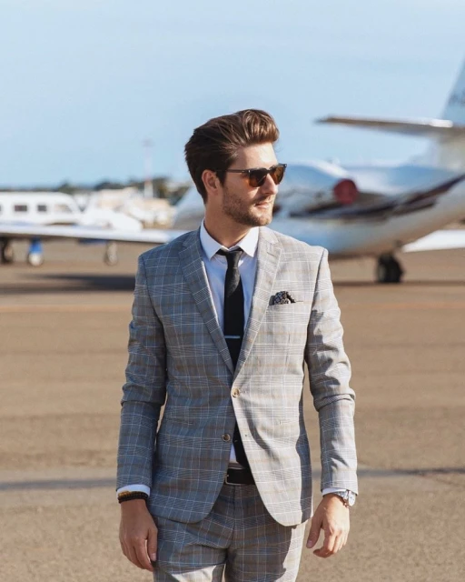 Stylish man in grey suit walks on airport tarmac with private jet in background, wearing sunglasses on a sunny day.
