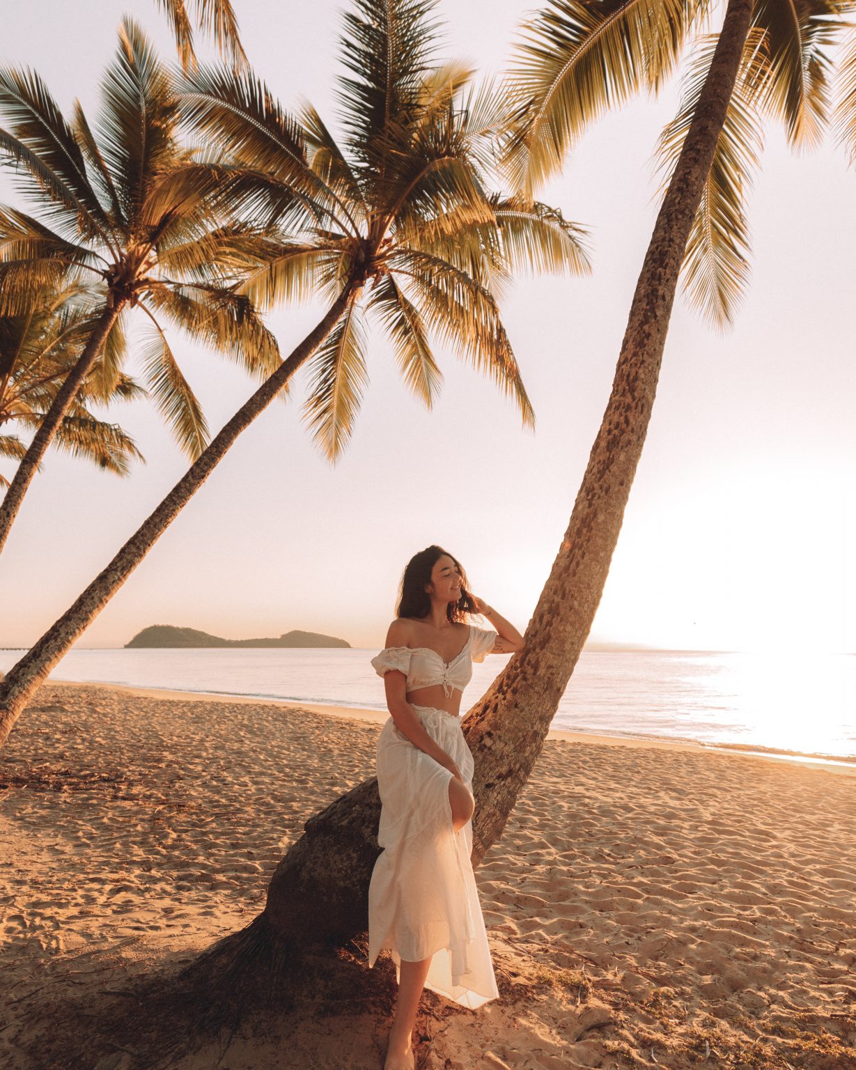 A woman in a white dress poses on a sandy beach by palm trees, bathed in golden sunlight with the ocean and a distant island visible.
