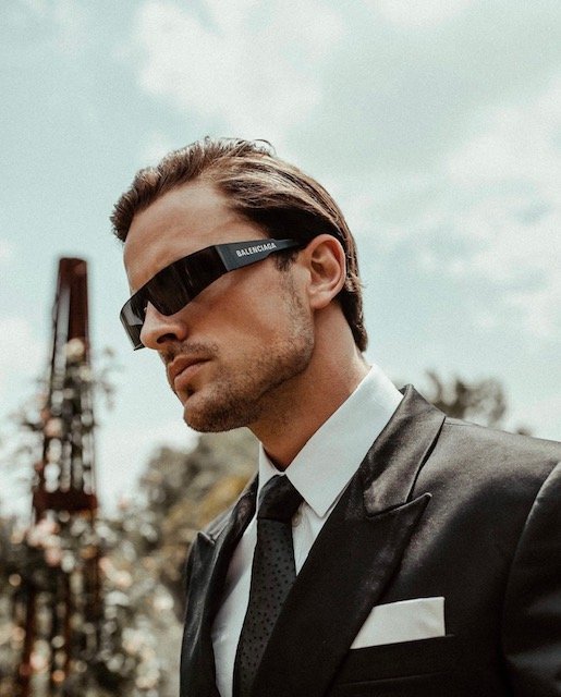 Portrait of an adult male model wearing a black suit, tie, and designer sunglasses, with a sky and floral background, close-up shot.
