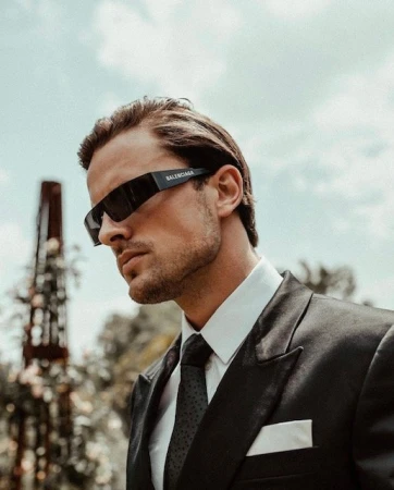 Portrait of an adult male model wearing a black suit, tie, and designer sunglasses, with a sky and floral background, close-up shot.