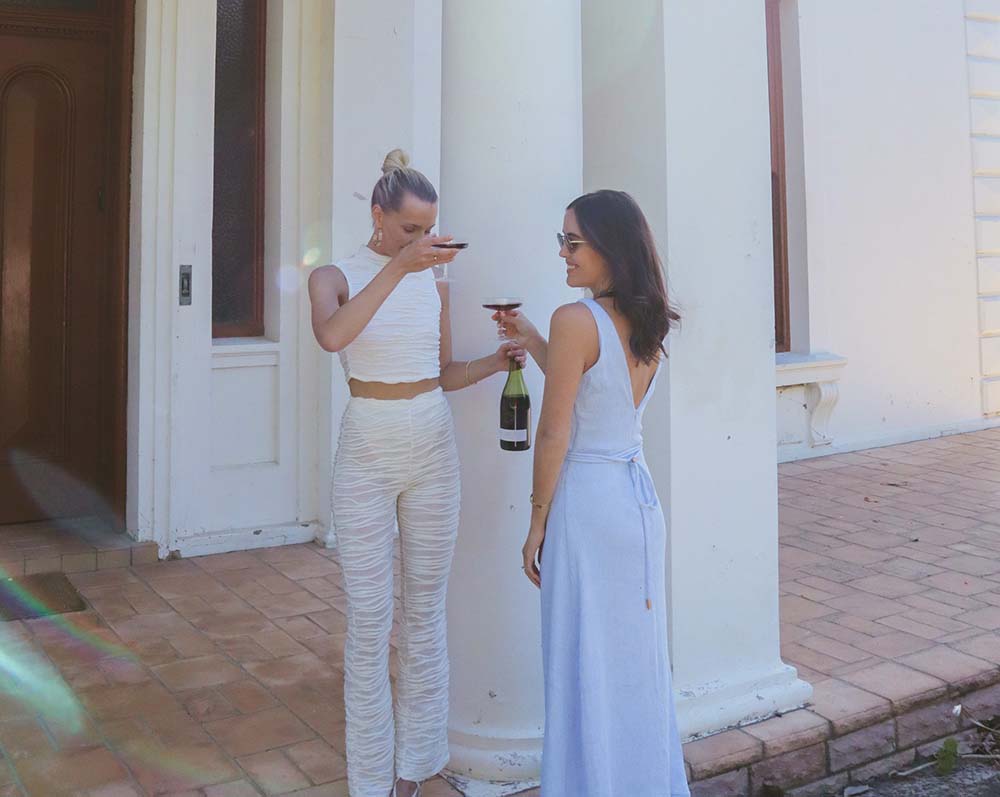 Two women in elegant attire share a bottle of red wine outside of a white building with prominent columns on a sunny day.