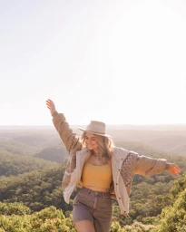 Young woman wearing a wide-brimmed hat and patterned jacket with arms raised outdoors against a valley view