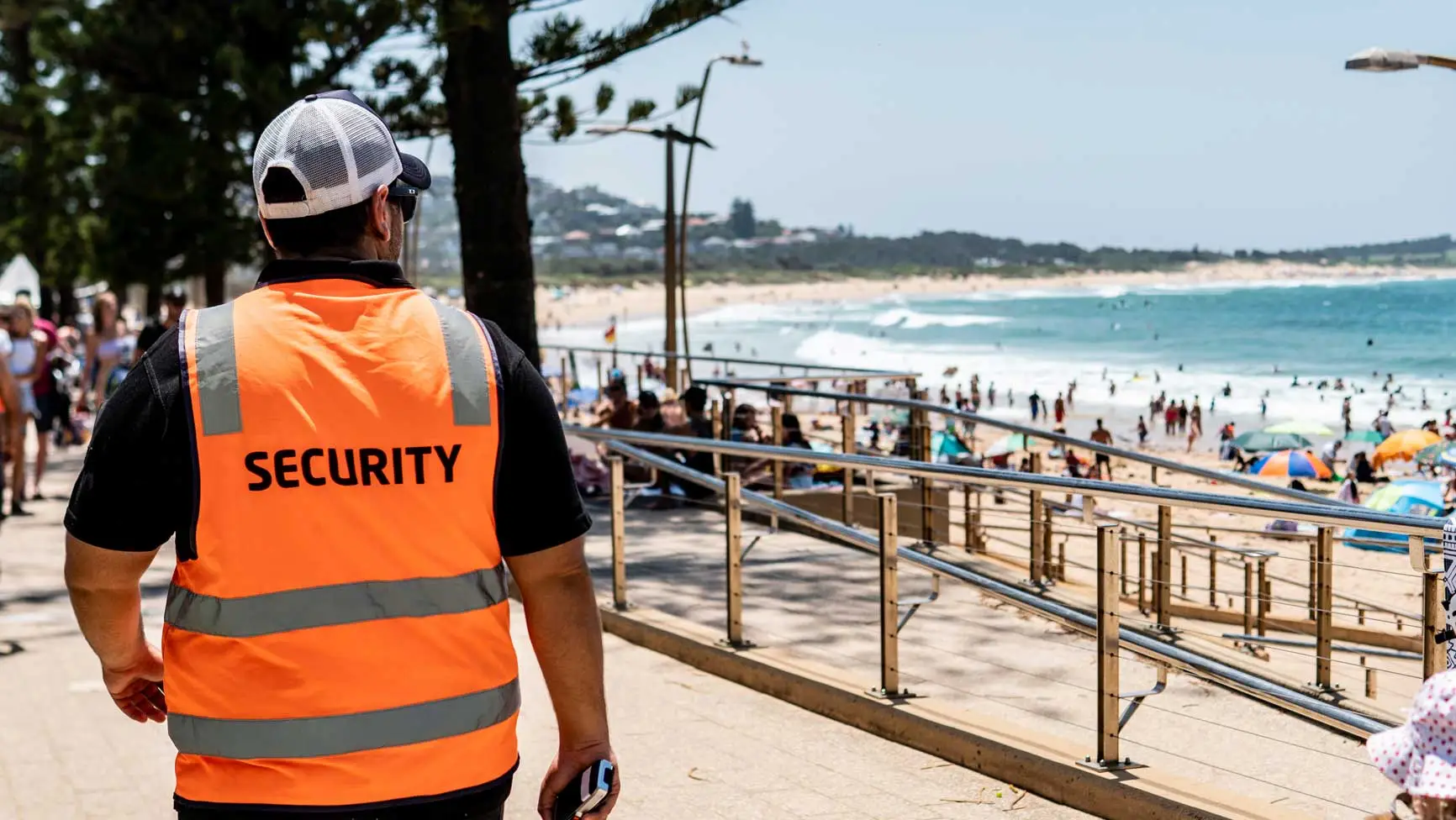 Security guard in orange vest patrolling a sunny beach with crowds, blue waves, and a clear sky in the background.