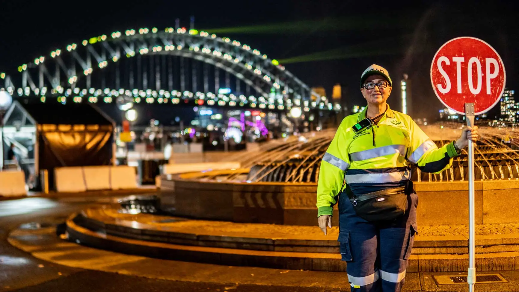 Adult female worker in a yellow safety jacket holding a stop sign with the Sydney Harbour Bridge in the background at night.