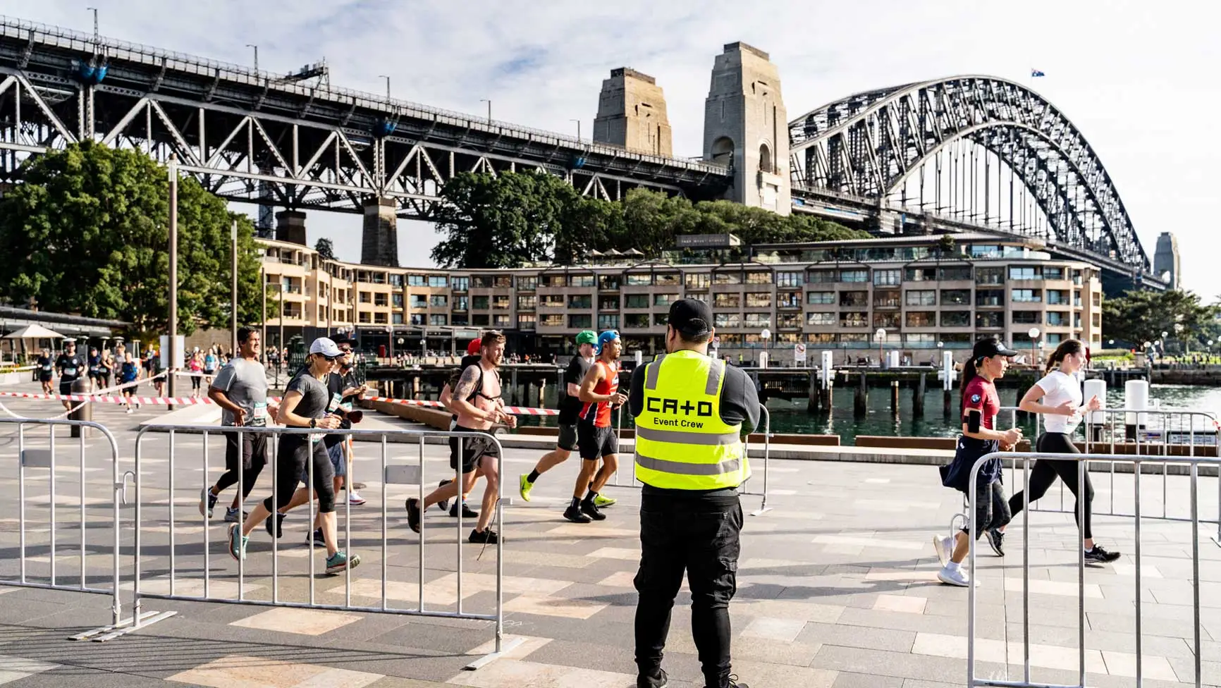 Sydney, Australia, daytime shot of a group of runners near the harbor with a security guard in a high-visibility vest in the foreground.