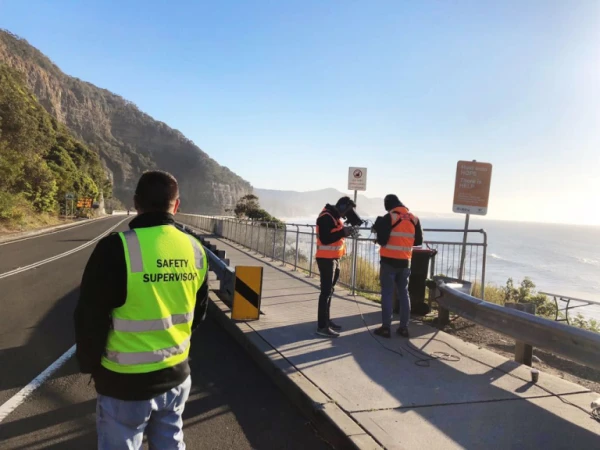 Three people in safety vests stand near a coastal road and the ocean, with a mountain in the background on a sunny day.