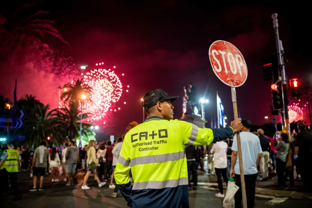 Traffic controller manages crowd with stop sign during a vibrant fireworks display at night in an outdoor, urban setting.