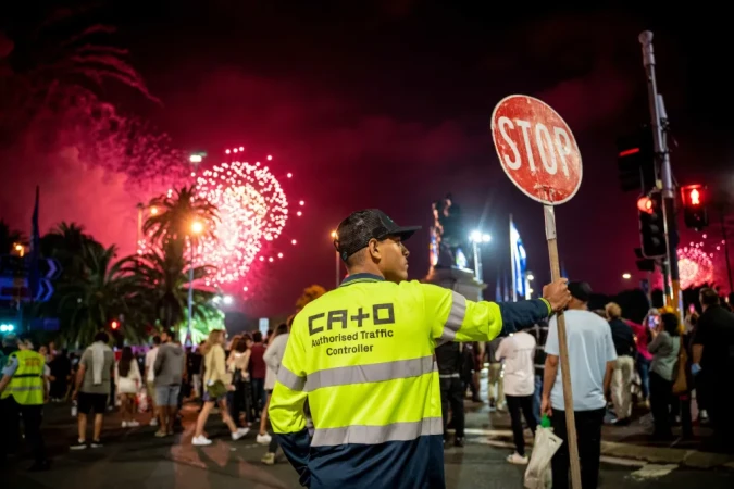 Traffic controller manages crowd with stop sign during a vibrant fireworks display at night in an outdoor, urban setting.