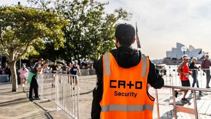 Security guard in orange vest stands near marathon race with Sydney Opera House in the background on a sunny day; tree line, city.