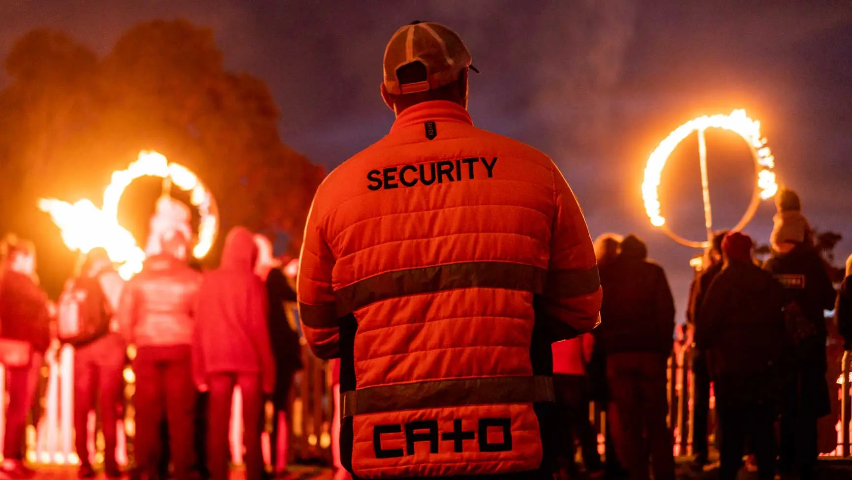Back view of security guard in orange jacket, patrolling a nighttime outdoor event with fire rings and a crowd of people in the background.