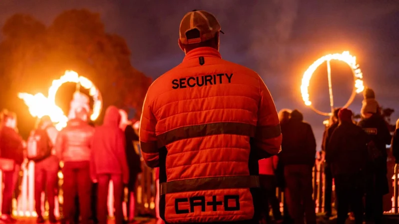 Back view of security guard in orange jacket, patrolling a nighttime outdoor event with fire rings and a crowd of people in the background.