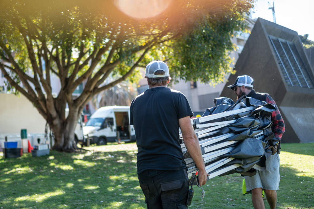 Two men carry folded tables outdoors on a sunny day with a tree and building in the background, capturing a moment of outdoor event preparation.