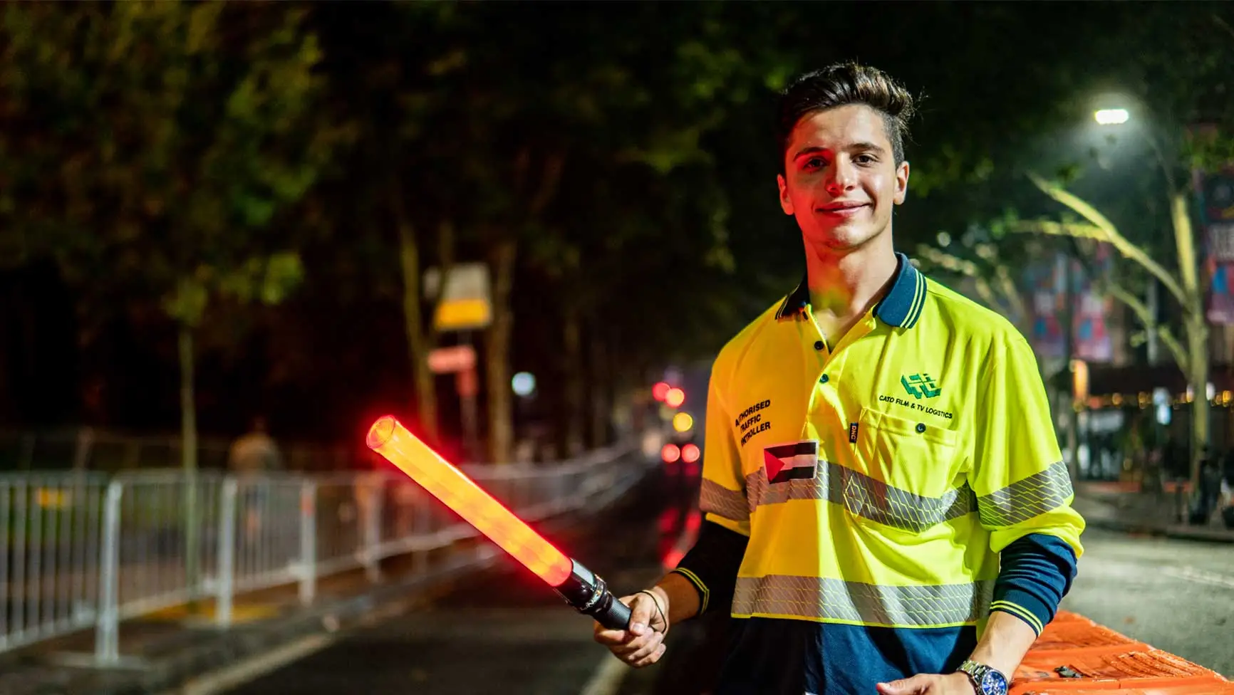 Adult man in yellow safety vest holding illuminated traffic wand on city street at night, smiling and looking toward the camera.
