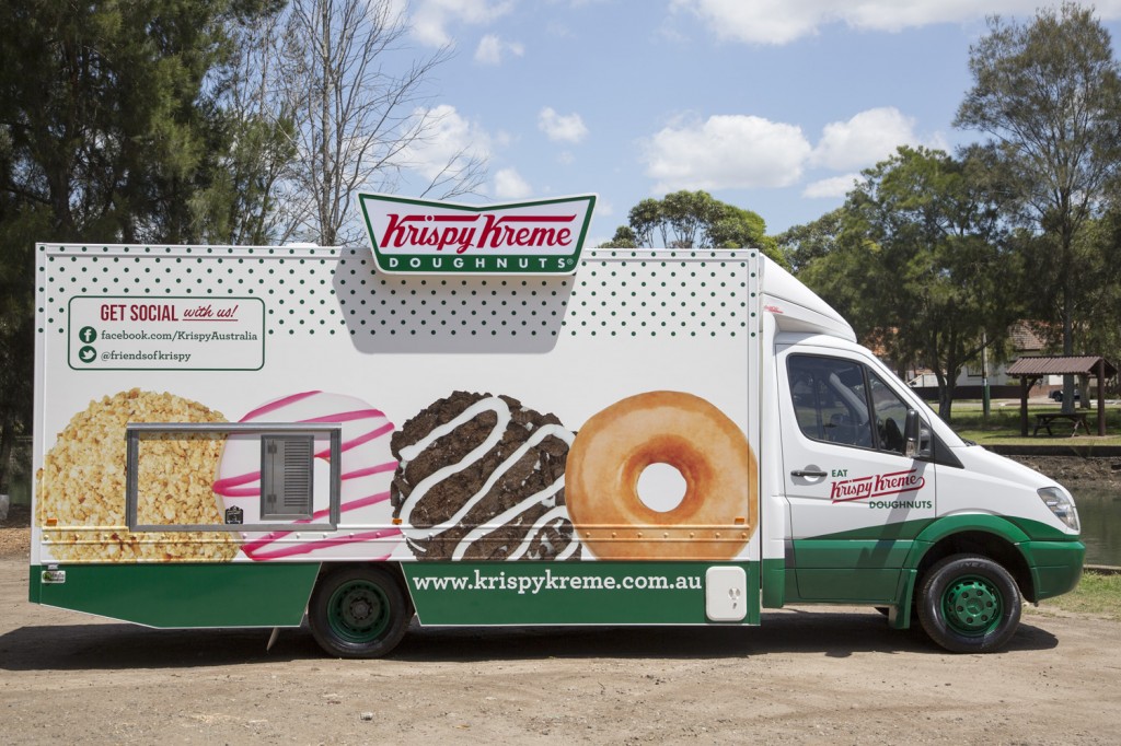 Krispy Kreme doughnuts food truck featuring branded logos and doughnut graphics parked outdoors on a sunny day near trees.
