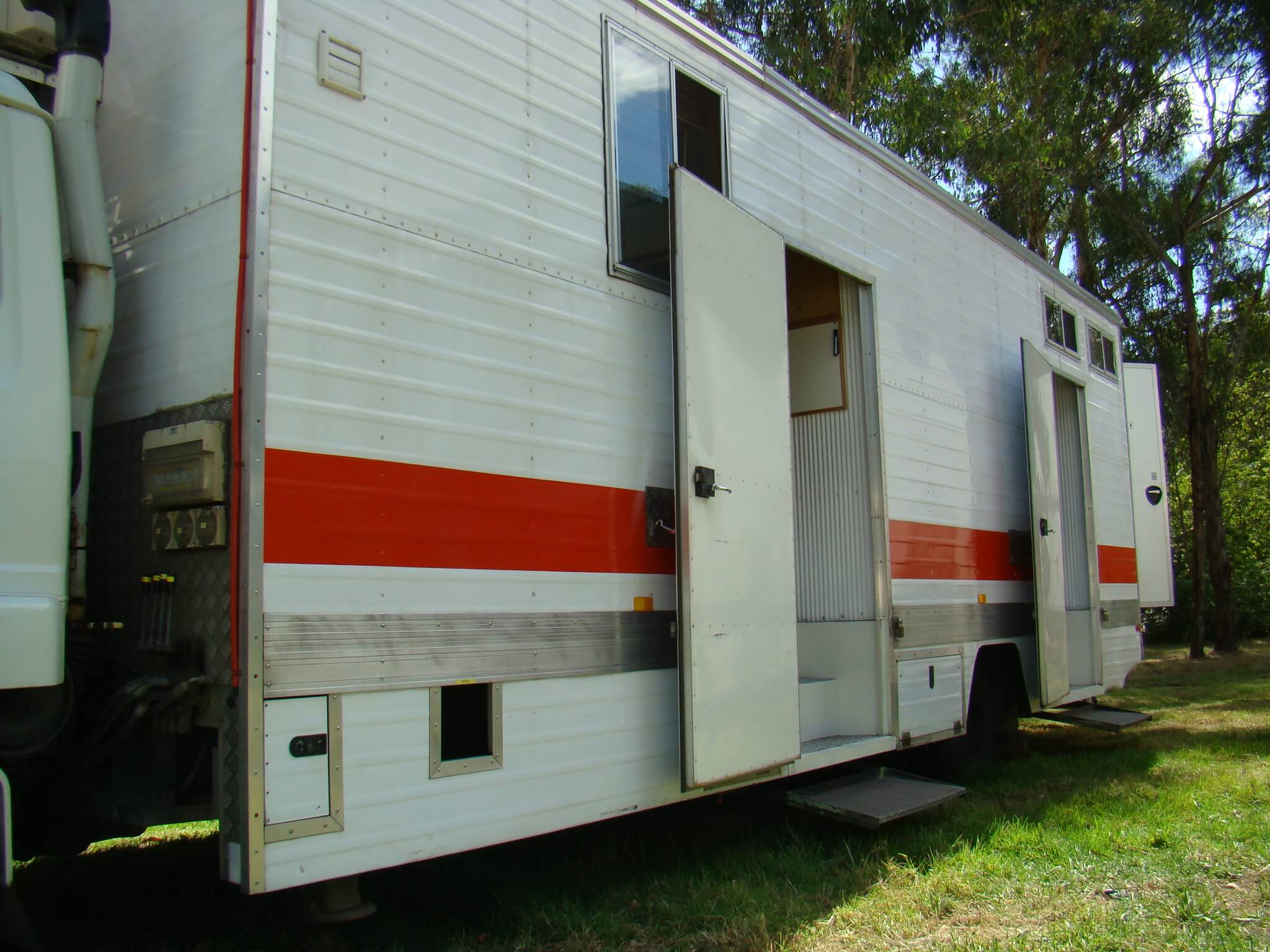 Long, white recreational vehicle with open doors parked on grassy land near trees, showcasing mobile living in a natural setting.