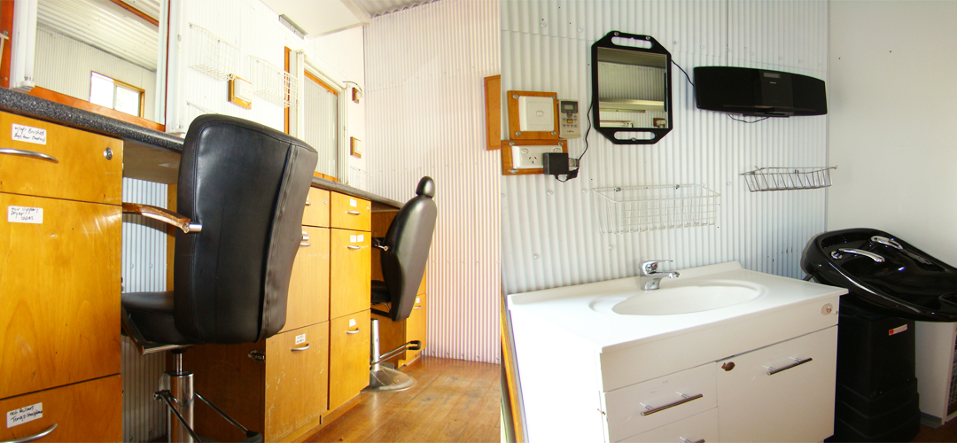 Interior of a hair salon featuring two black styling chairs, wooden cabinets, a sink, and a shampoo basin against white corrugated walls.