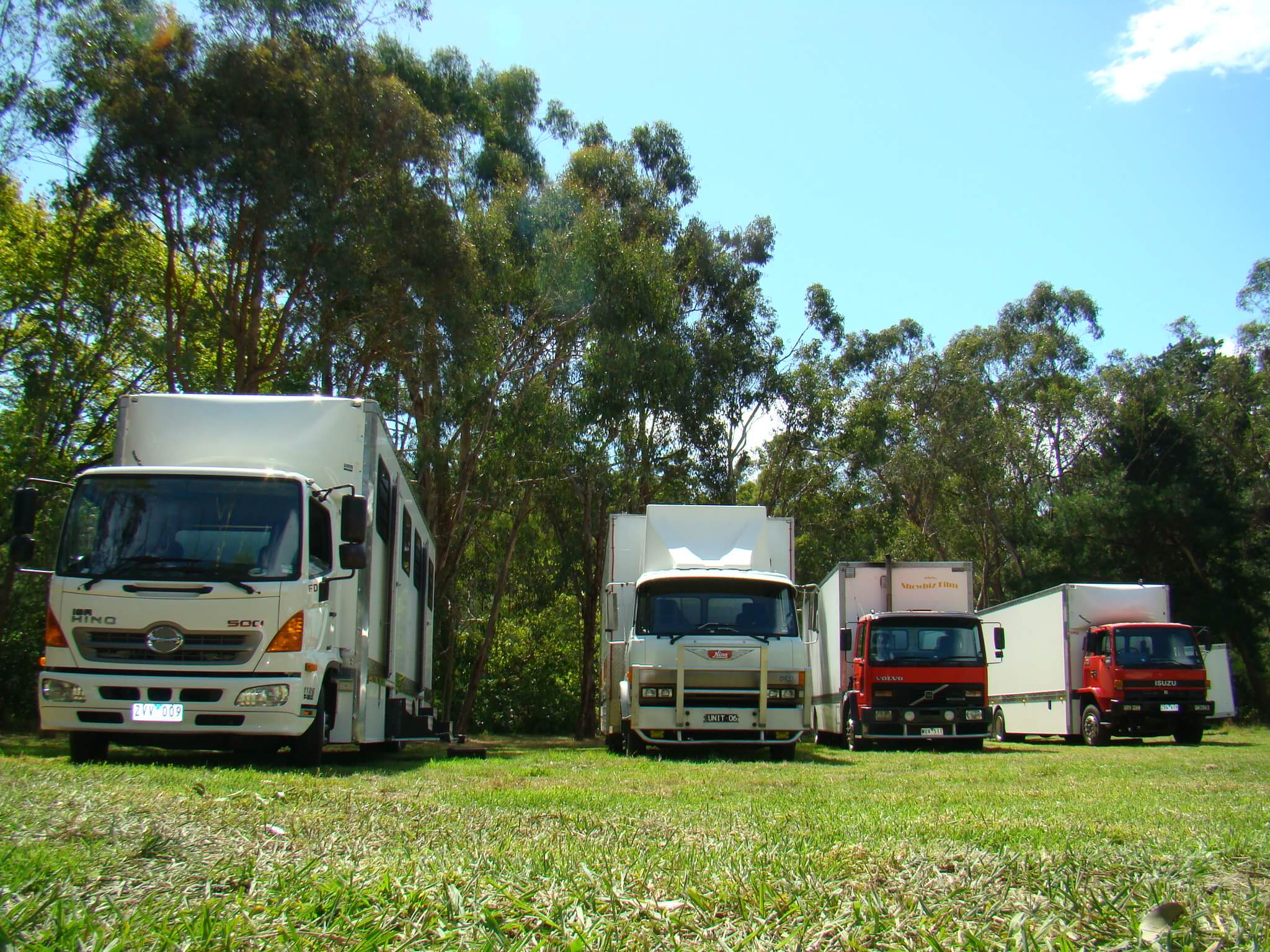 Several white and red trucks are parked in a grassy field surrounded by trees under a clear blue sky, creating a bright outdoor scene.
