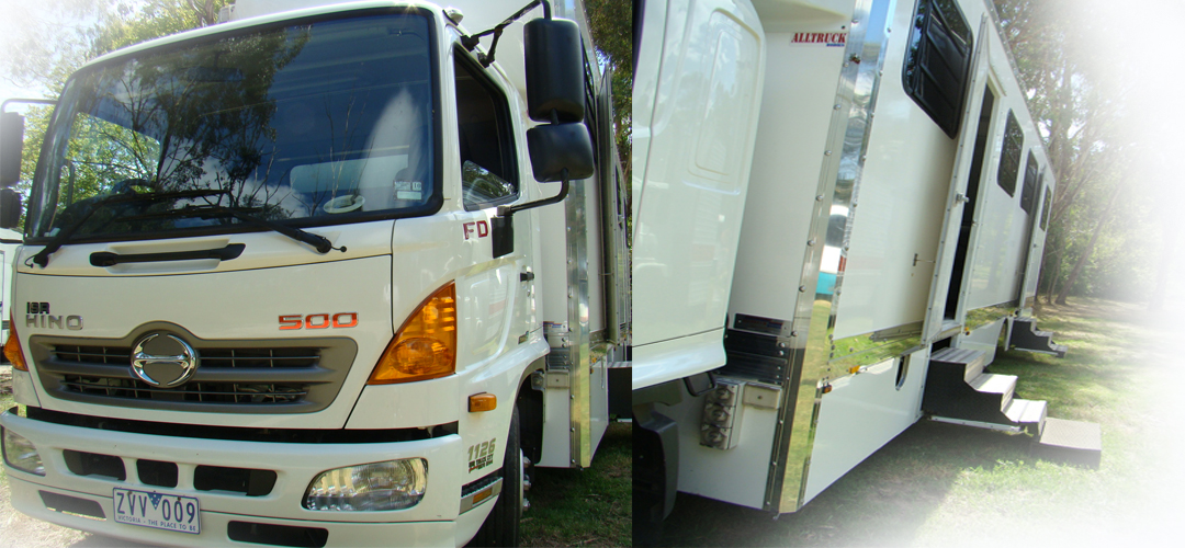A white Hino FD 500 truck with a modified cabin and steps on a grassy surface. The modified vehicle stands out against a natural backdrop.