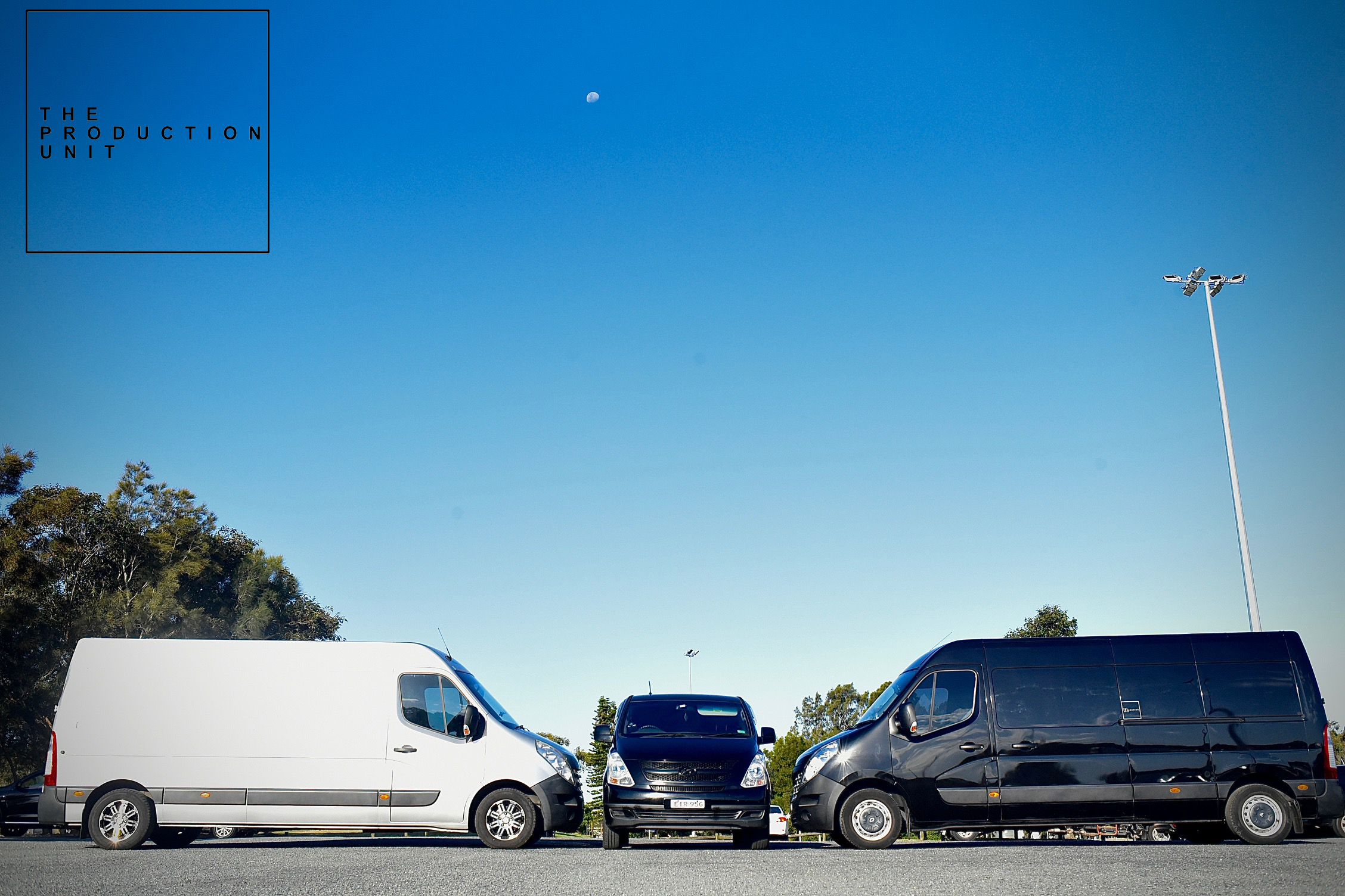 Three commercial vans are parked side by side on asphalt under a clear blue sky with trees and a tall light pole in the background.