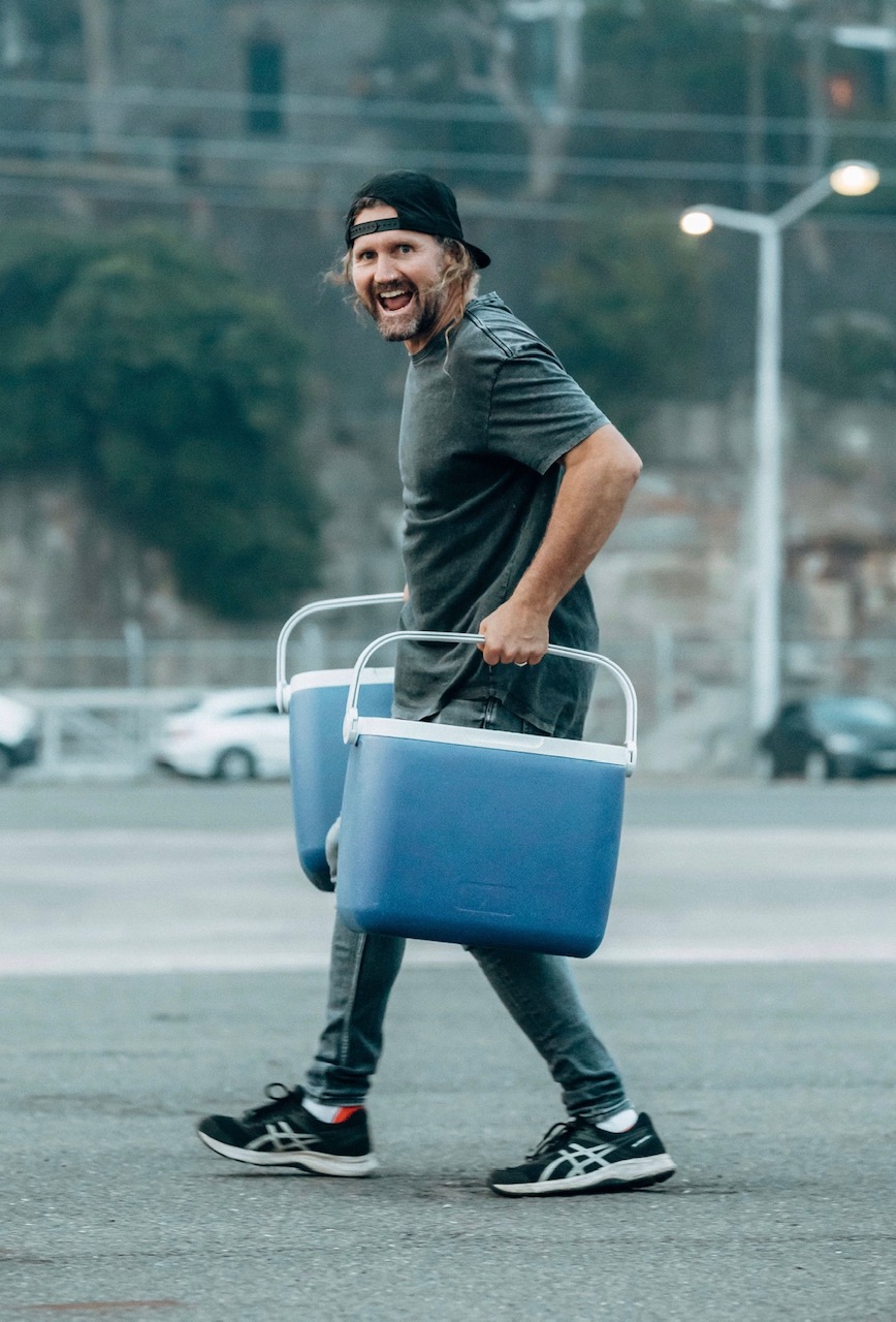 Adult man in a baseball cap carries two blue coolers while walking on a city street, looking toward the camera with an open mouth.
