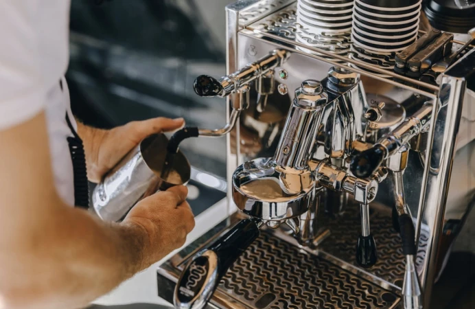 Close-up of hands using a professional espresso machine to steam milk into a metal pitcher in a cafe environment, creating artisan coffee.
