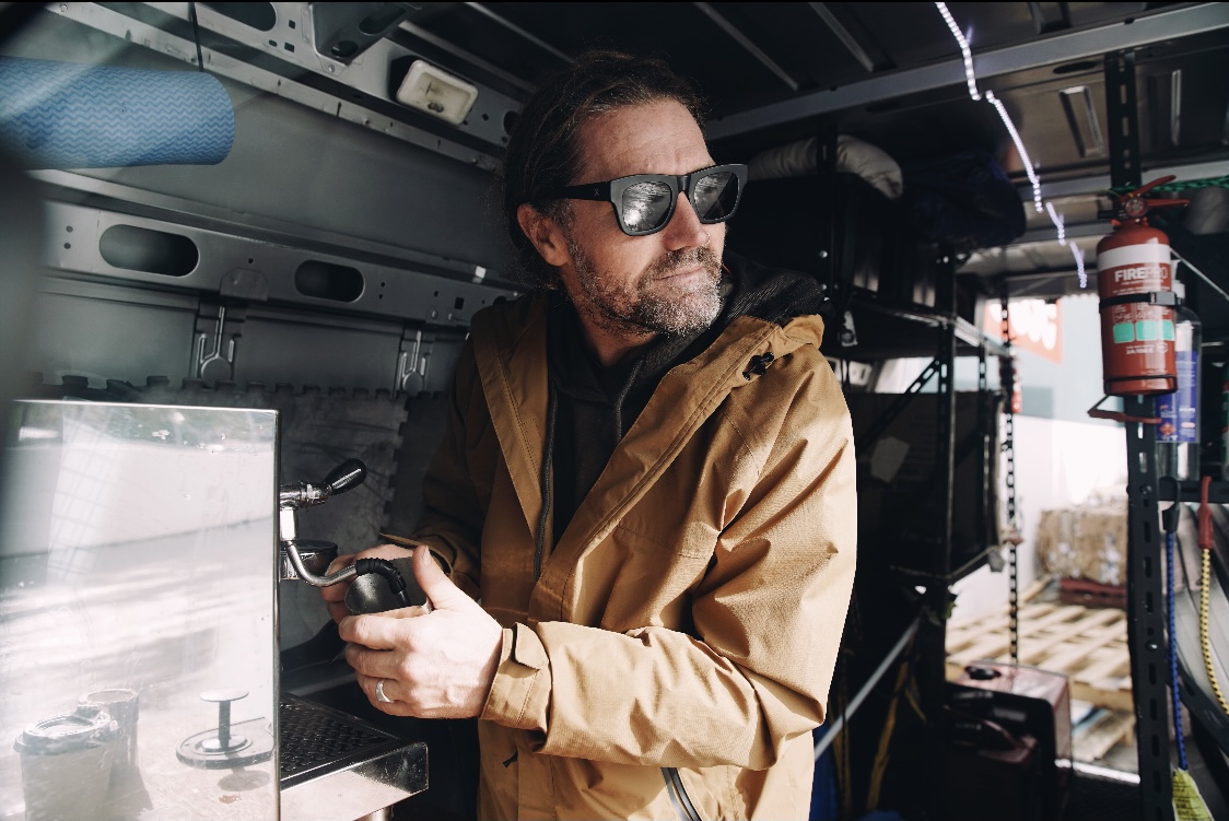 An adult male with sunglasses operates an espresso machine inside of a van. Shelves and equipment fill the interior of the vehicle.