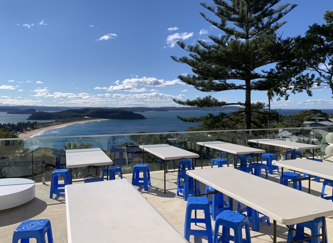 Scenic outdoor venue with white tables and blue stools overlooking a beautiful ocean view with clear skies and lush green landscapes.
