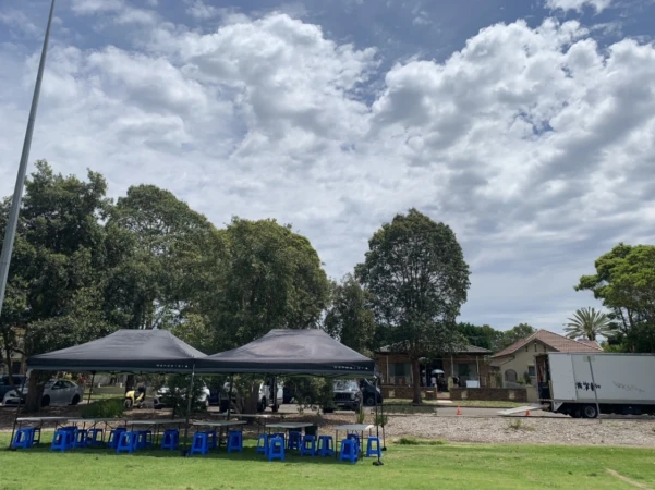 Outdoor setup with canopies, tables, and blue plastic chairs on a grassy field, with trees, a building, and cloudy skies in the background.
