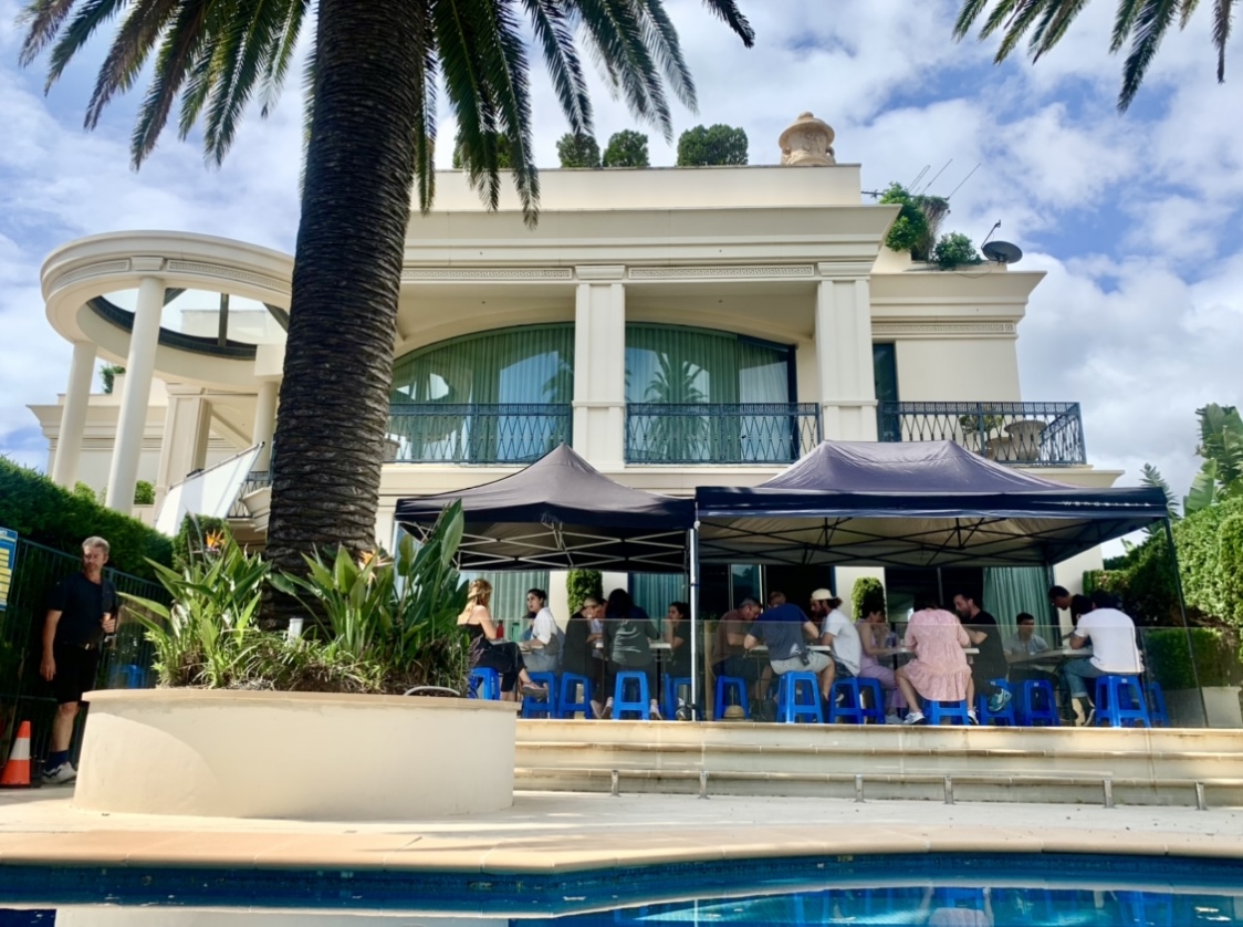 Outdoor gathering by the pool with multiple people seated under tents near a large house surrounded by palm trees on a sunny day.