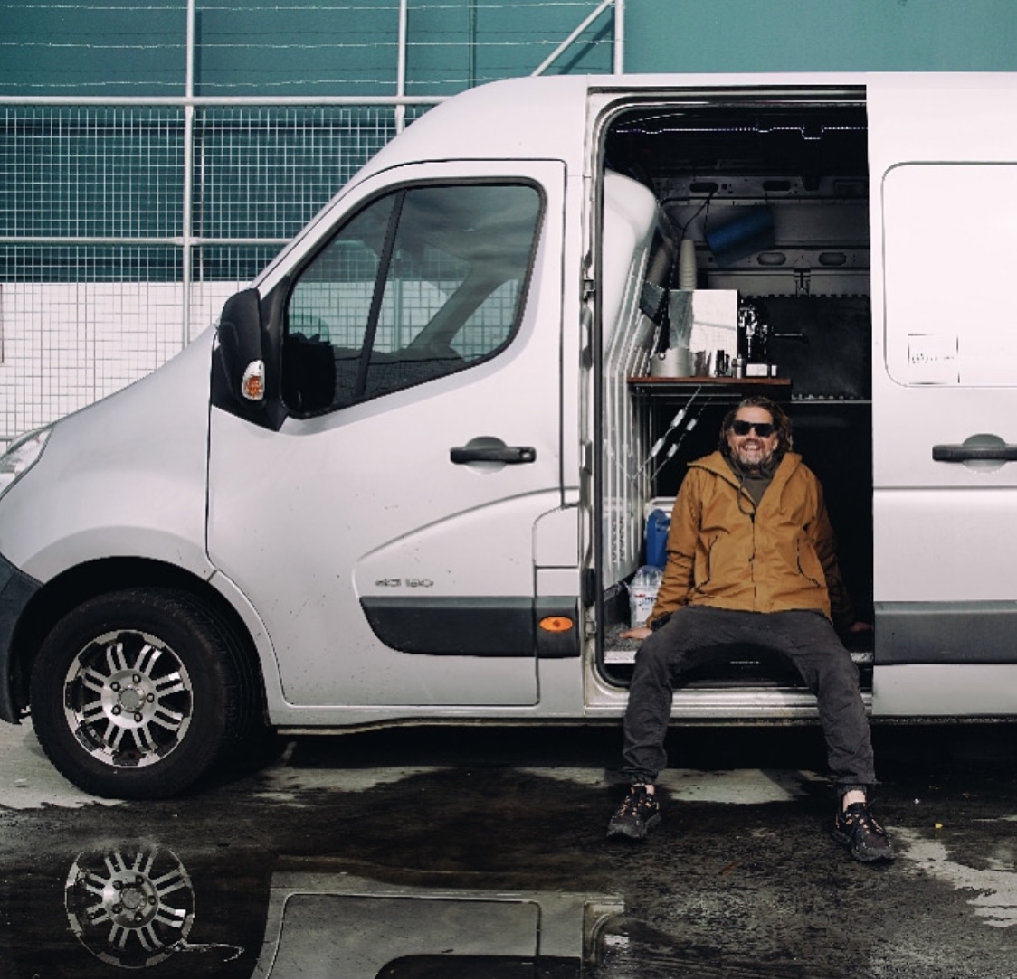 Adult male with sunglasses sits in the doorway of a silver van with cargo area visible, parked outdoors on a wet surface.