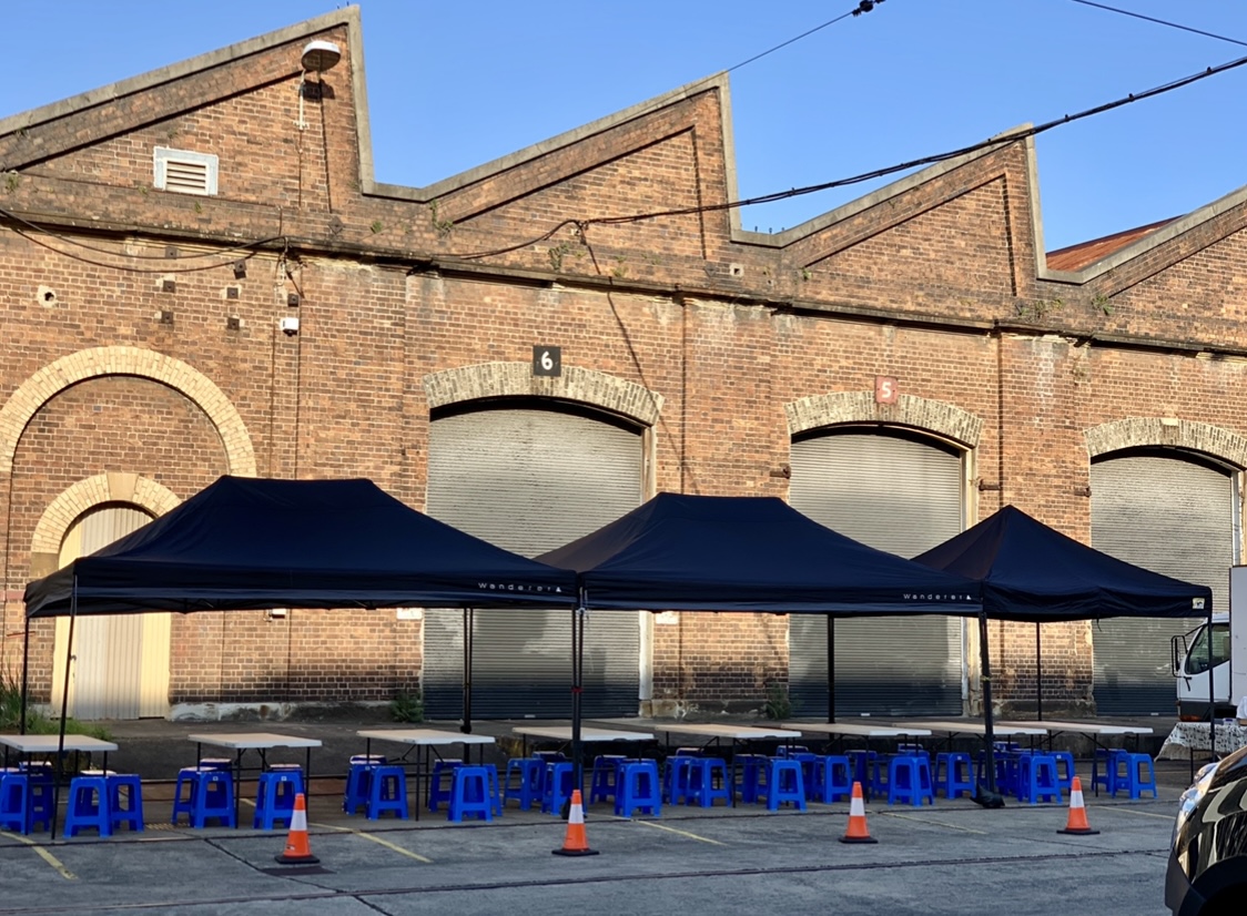 Outdoor event setup with tables and blue chairs under canopies in front of a brick building on a sunny day, creating an inviting atmosphere.