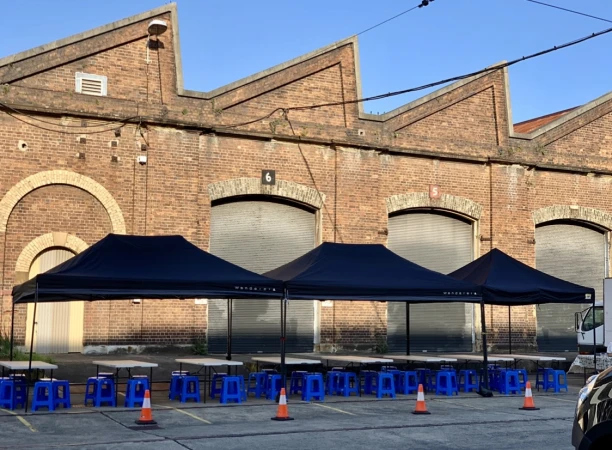 Outdoor event setup with tables and blue chairs under canopies in front of a brick building on a sunny day, creating an inviting atmosphere.