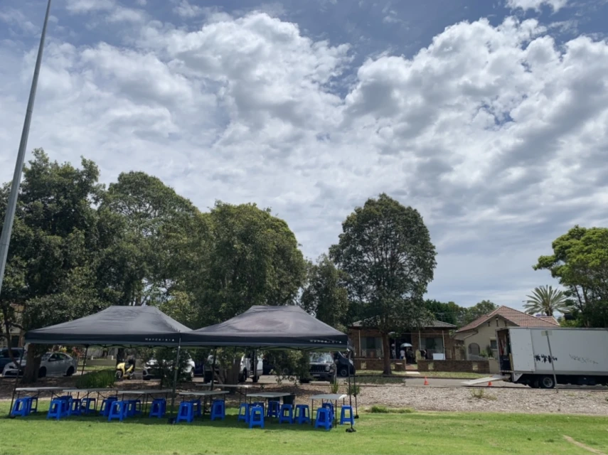 Production tents and blue plastic stools set up on grass in Sydney under a partly cloudy sky with trees and buildings in the background.