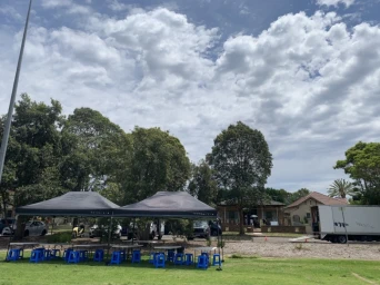 Production tents and blue plastic stools set up on grass in Sydney under a partly cloudy sky with trees and buildings in the background.