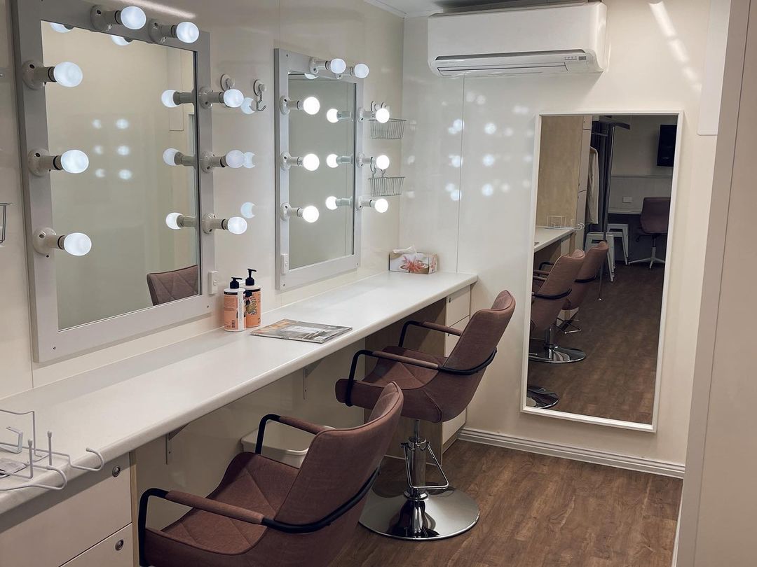 Interior of a hair and makeup salon with vanity mirrors, chairs, and counters. White walls, brown floors, and good lighting create a clean.