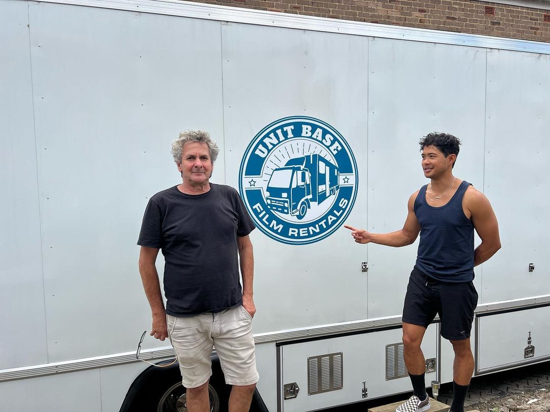 Two men stand in front of a white film unit trailer with the logo UNIT BASE FILM RENTALS, outdoors on a sunny day.