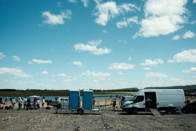 Outdoor scene of a film or video shoot with crew, equipment, portable toilets, and a van under a partly cloudy sky near a body of water.