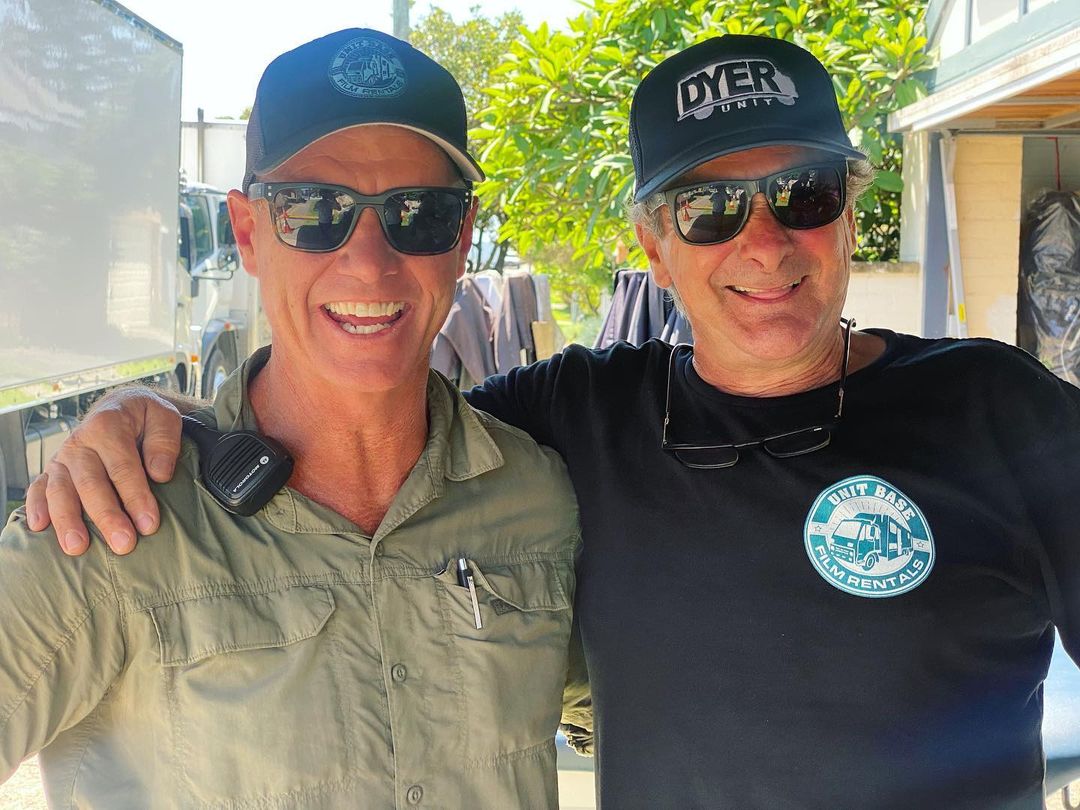 Two adult men wearing hats and sunglasses stand close together and smile in an outdoor setting with a vehicle and trees in the background.