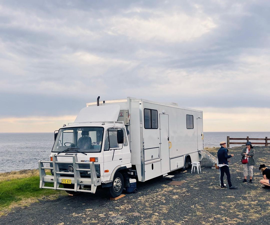 A white camper van parked on a bluff overlooking the ocean under an overcast sky, with a group of people standing nearby.
