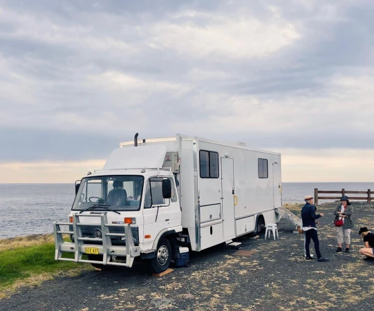 A white camper van parked on a bluff overlooking the ocean under an overcast sky, with a group of people standing nearby.