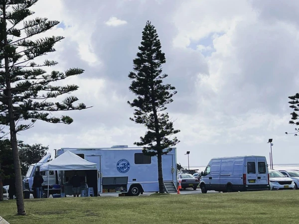 Mobile medical unit parked on a grassy area near a parking lot, with pine trees and a cloudy sky in the background. 