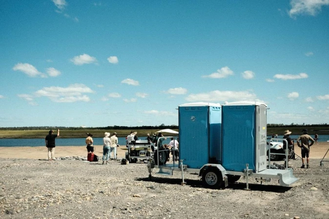 Outdoor film set with portable restrooms, crew, and equipment near a lake under a bright sky, capturing a behind-the-scenes production scene.