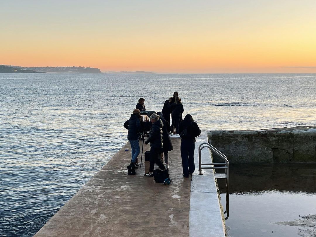 Film crew with cameras gathers on a pier by the ocean at sunset, capturing the tranquil seascape in a behind-the-scenes shot.