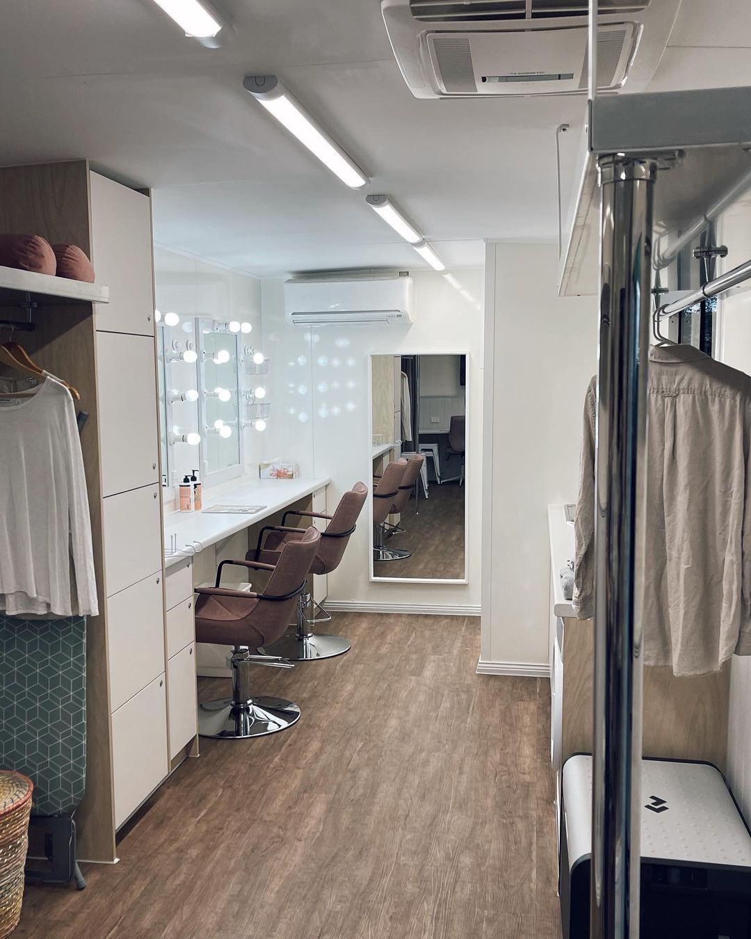 Interior shot of a modern makeup room featuring several makeup stations, chairs, lighting fixtures, and minimalist decor.