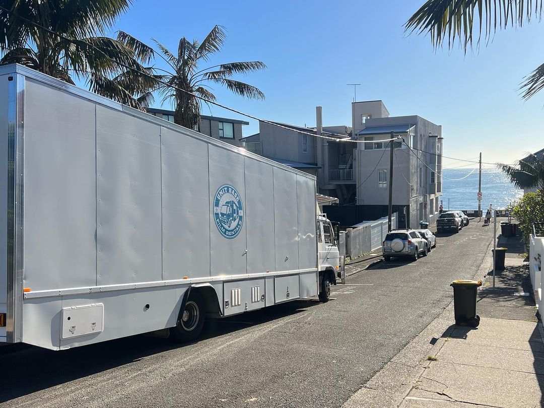 Commercial box truck parked on a residential street near the ocean, with modern houses, palm trees, blue sky, and parked cars.