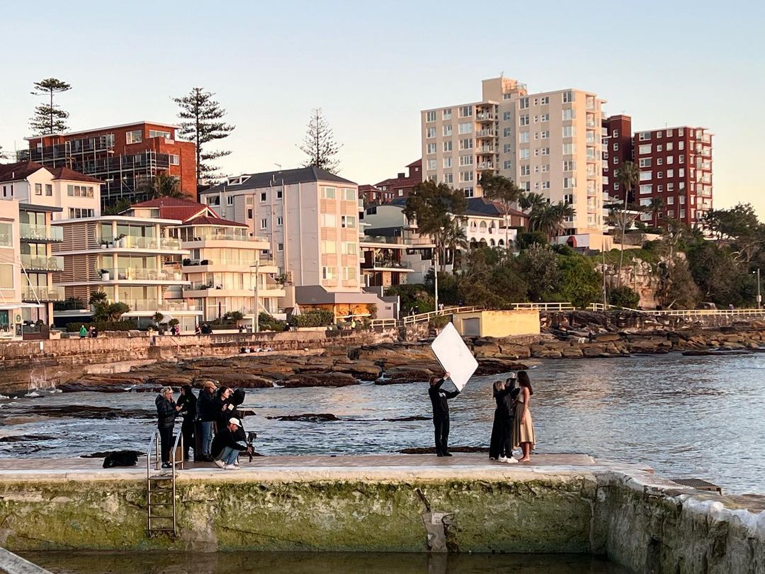 Outdoor fashion shoot by the ocean in a saltwater pool at sunset; models pose with a crew using a light reflector by the sea.